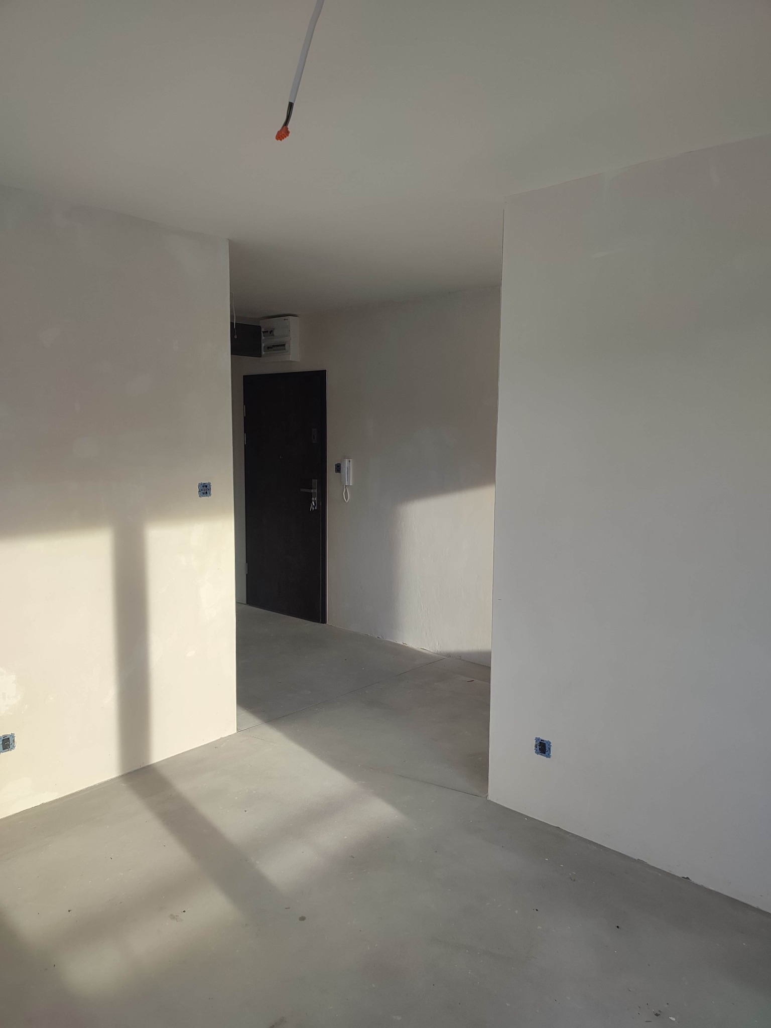 Wnętrze w stanie deweloperskim z gładkimi, pomalowanymi na biało ścianami, surową betonową podłogą, widocznymi puszkami elektrycznymi i wystającymi kablami z sufitu, czarne drzwi wejściowe w głębi.