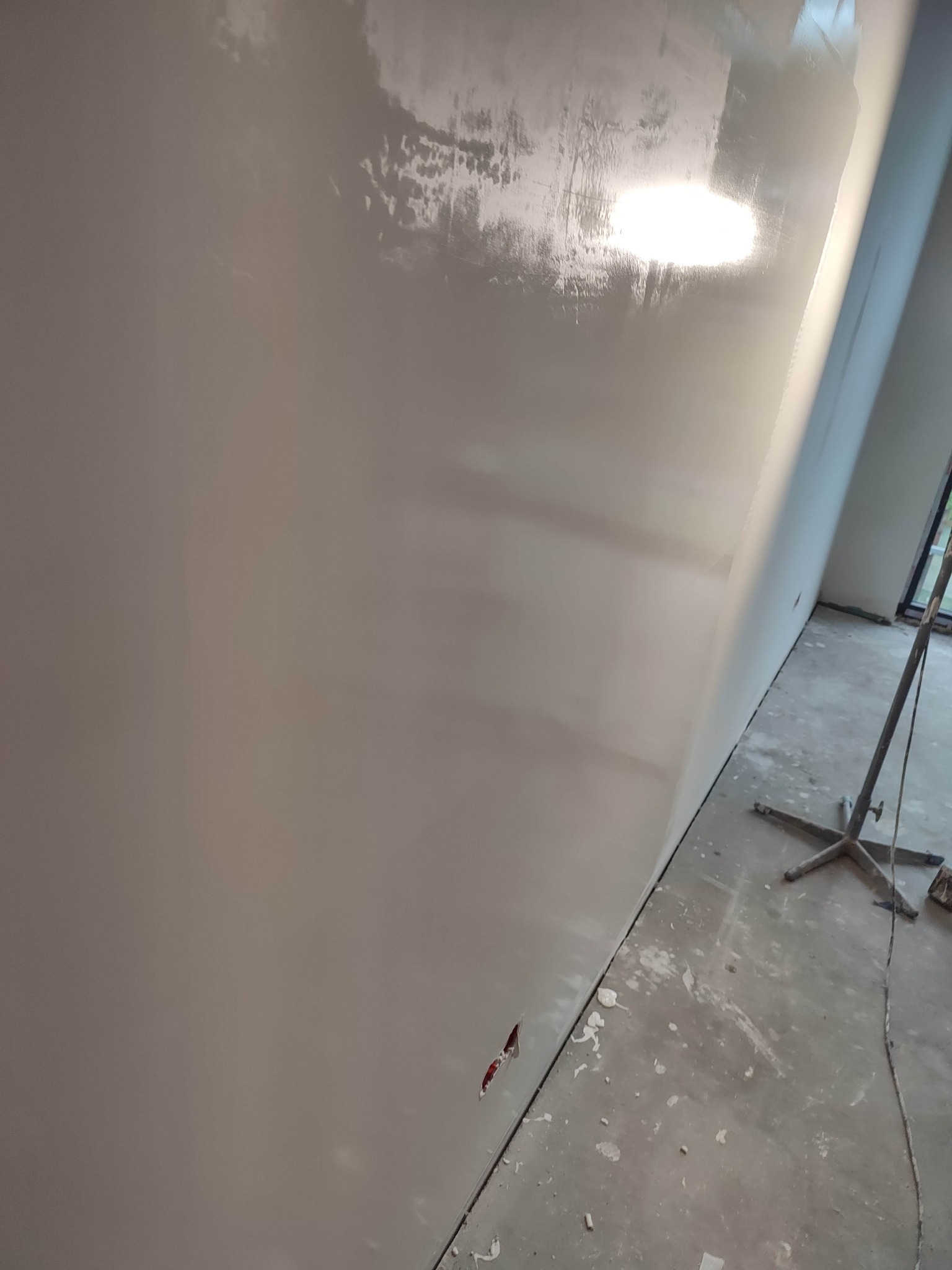 Świeżo pomalowana, błyszcząca ściana w odcieniach szarości, z widocznymi smugami farby i niedoskonałościami wykończenia, w pomieszczeniu w trakcie remontu z zabrudzonym betonowym podłożem...