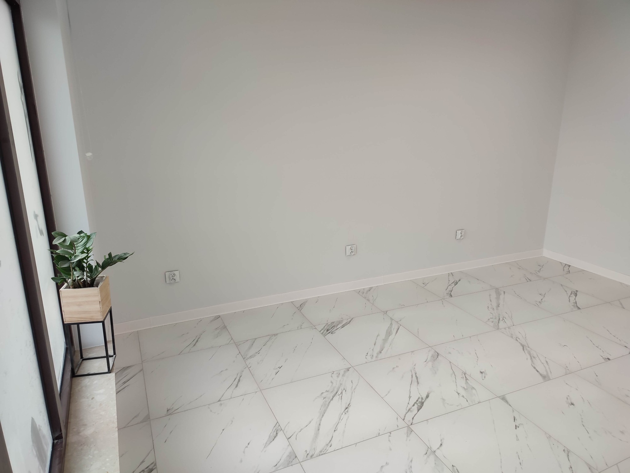 Puste, jasne pomieszczenie z nową podłogą z płytek imitujących marmur, białe ściany z widocznymi gniazdkami elektrycznymi i rośliną doniczkową w drewnianej osłonce na metalowym stojaku przy oknie.