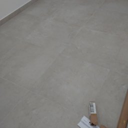 ANTEX - Wyłożona jasnymi, kwadratowymi płytkami podłoga w pustym pomieszczeniu, z widocznymi fragmentami kartonowego opakowania i dokumentu w rogu.