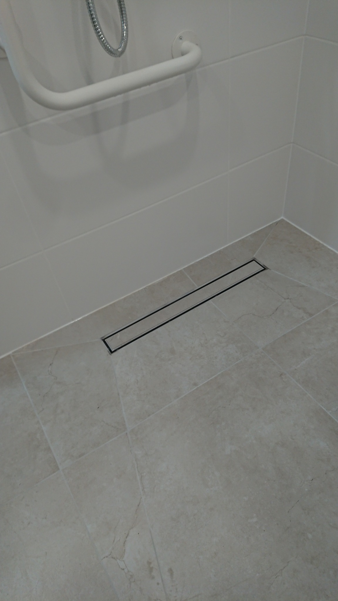 Beżowa, wykafelkowana podłoga prysznica z liniowym odpływem, uchwyt dla niepełnosprawnych i chromowana słuchawka prysznicowa.