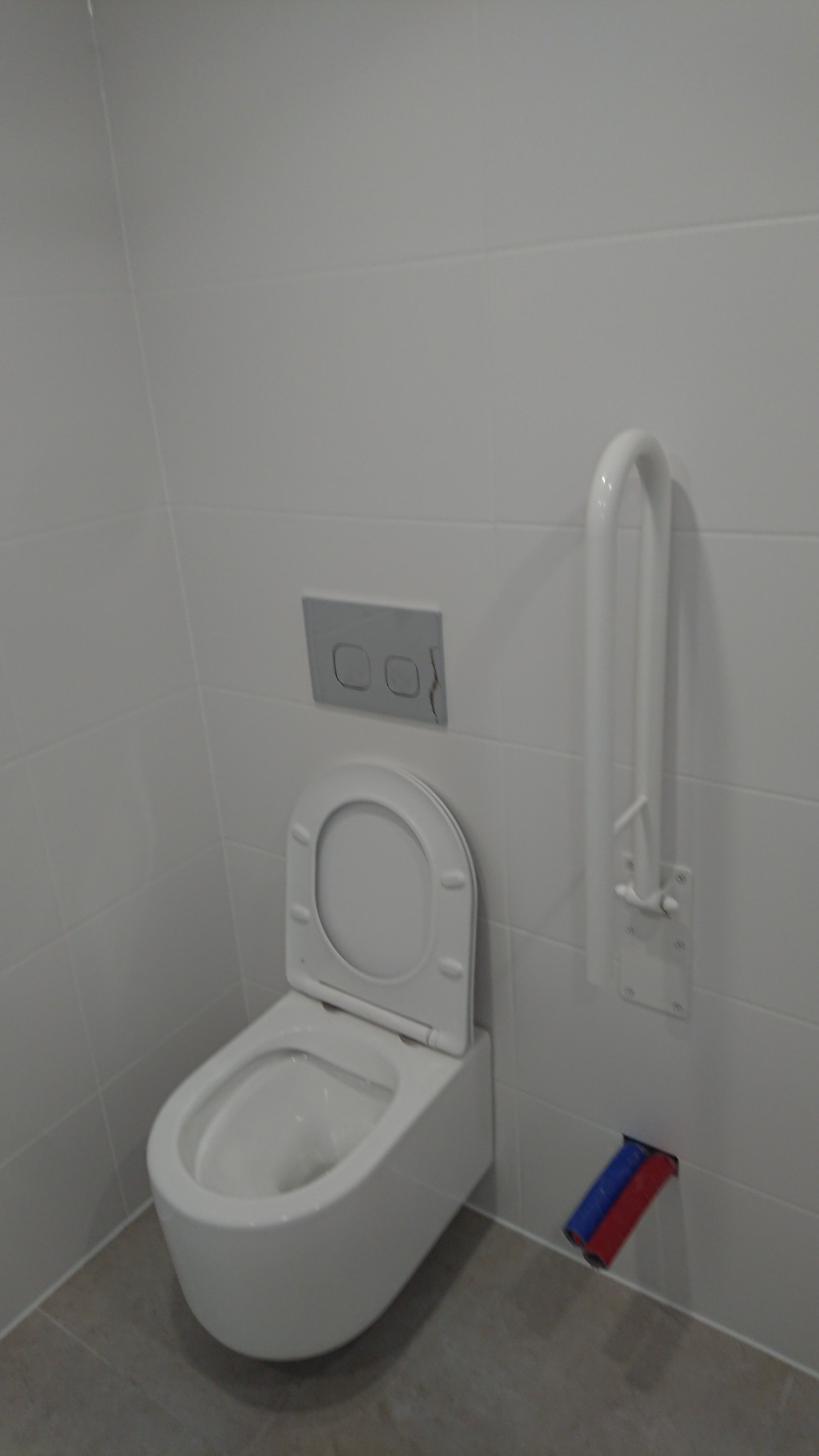 Biała podwieszana toaleta z otwartą klapą, przycisk spłuczki z pęknięciem i poręcz dla niepełnosprawnych na tle białych płytek, widoczne rury doprowadzające wodę.