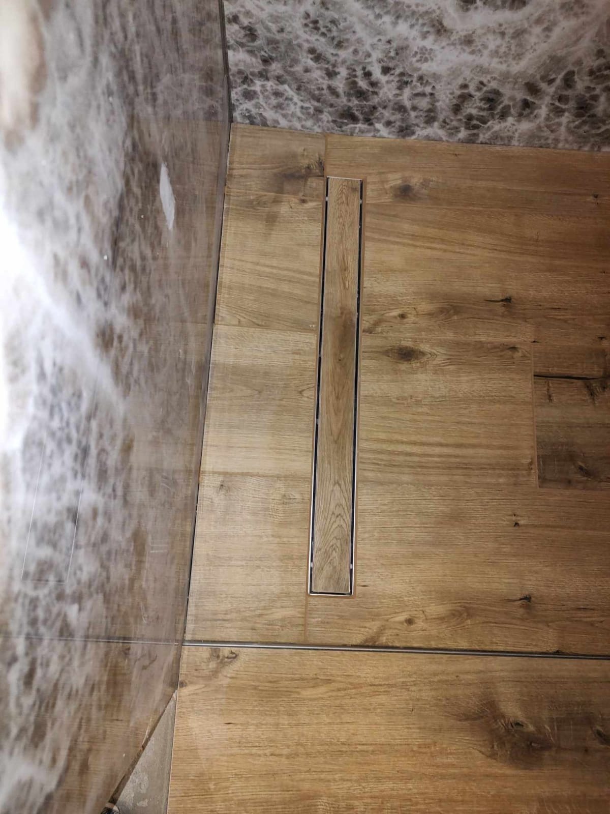 Szczegółowe ujęcie podłogi w łazience wyłożonej płytkami imitującymi drewno, z liniowym odpływem prysznicowym w metalowej ramie oraz fragmentem ściany pokrytej marmurkowymi płytkami.