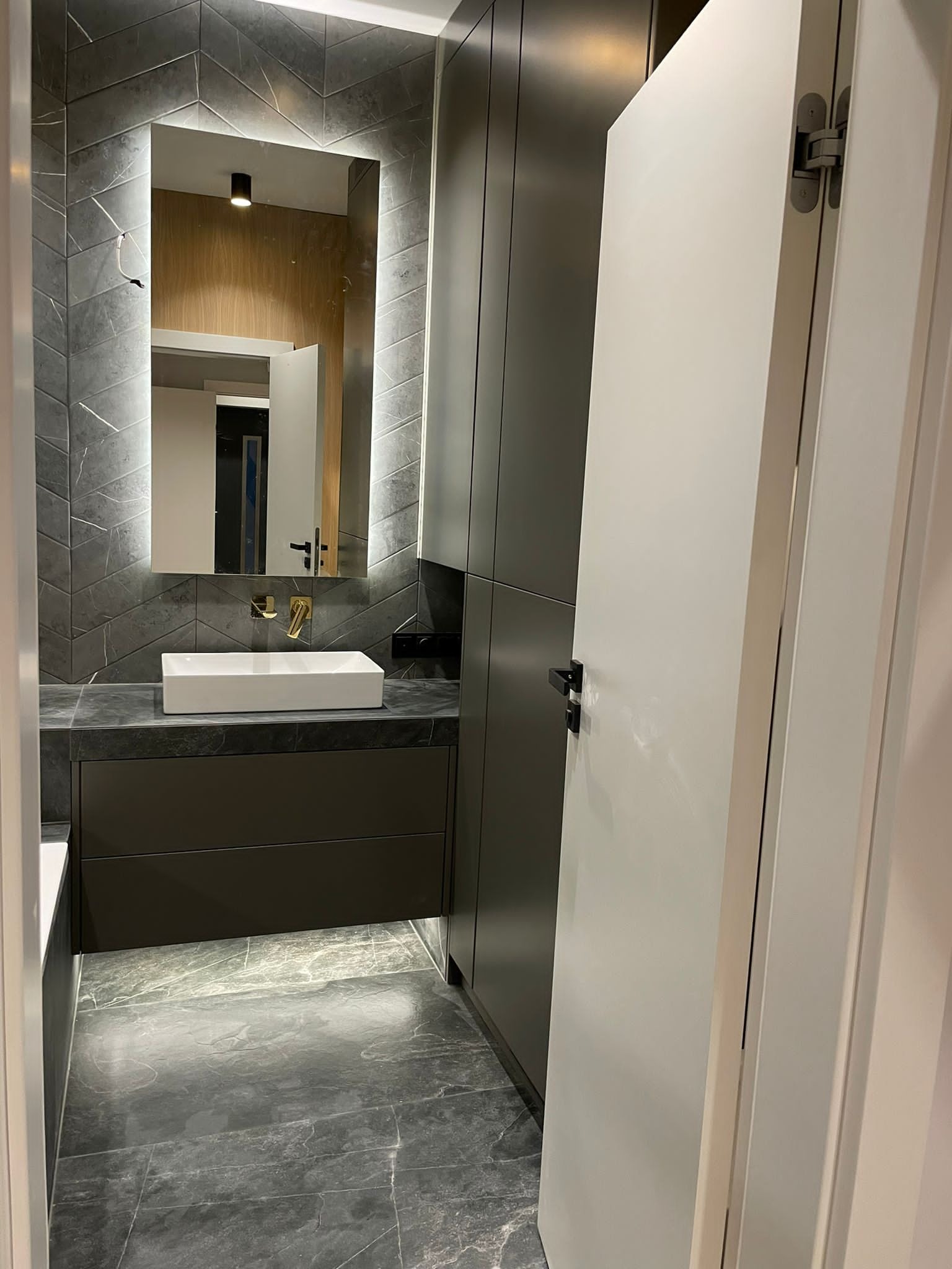 Nowoczesna łazienka z szarymi płytkami ułożonymi w jodełkę na ścianie, podświetlanym lustrem, białą umywalką nablatową, szarą szafką pod umywalką i marmurową podłogą, widoczne drzwi z ukrytymi...