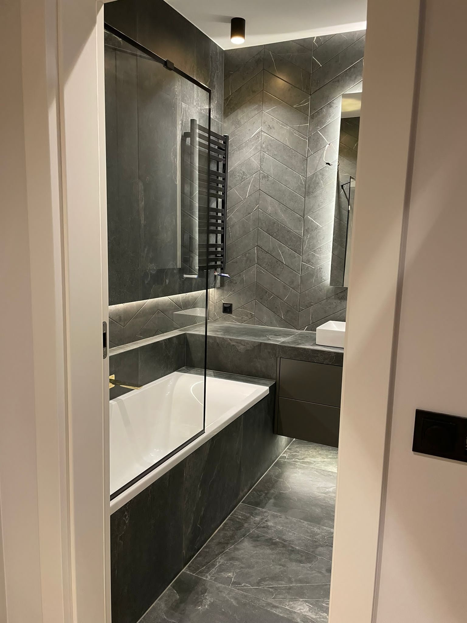 Nowoczesna łazienka w odcieniach szarości z płytkami ułożonymi w jodełkę, podświetlaną wanną z parawanem i grzejnikiem drabinkowym.