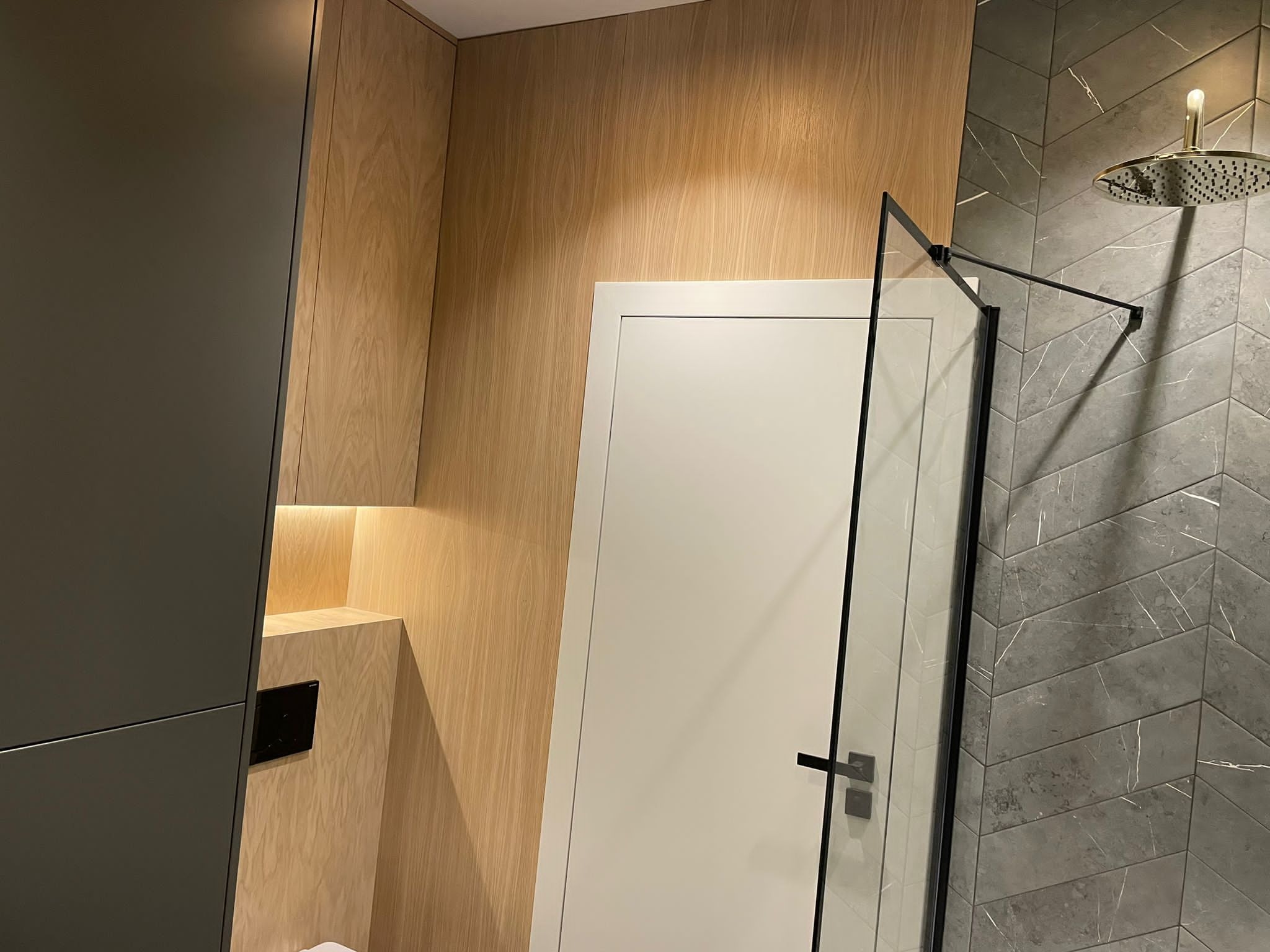 Nowoczesna łazienka z drewnianymi panelami, szarą mozaiką w jodełkę pod prysznicem z deszczownicą i minimalistyczną kabiną prysznicową z czarnymi profilami.
