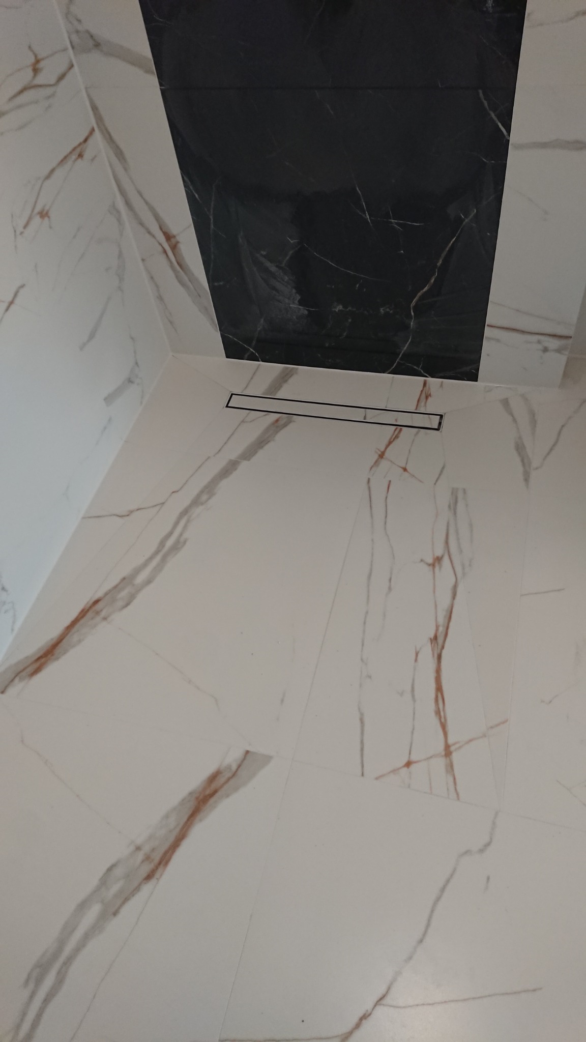 Wykończona kabina prysznicowa z odpływem liniowym, wyłożona białymi płytkami imitującymi marmur z brązowymi żyłami, połączona z czarną płytką na ścianie.