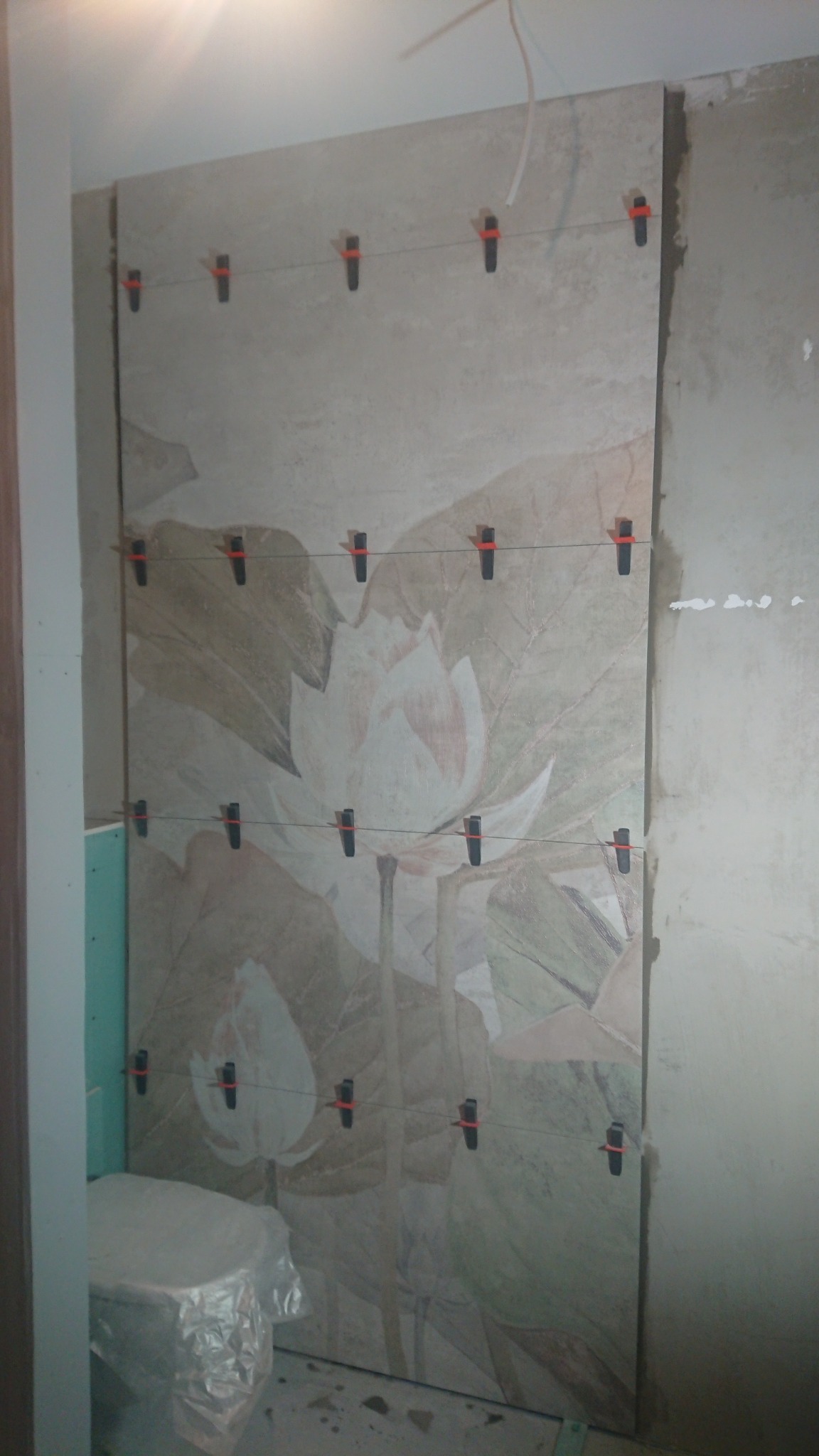 Ściana w trakcie układania płytek z motywem kwiatowym, z użyciem klipsów poziomujących, widoczne surowe ściany i zakryta folią toaleta.