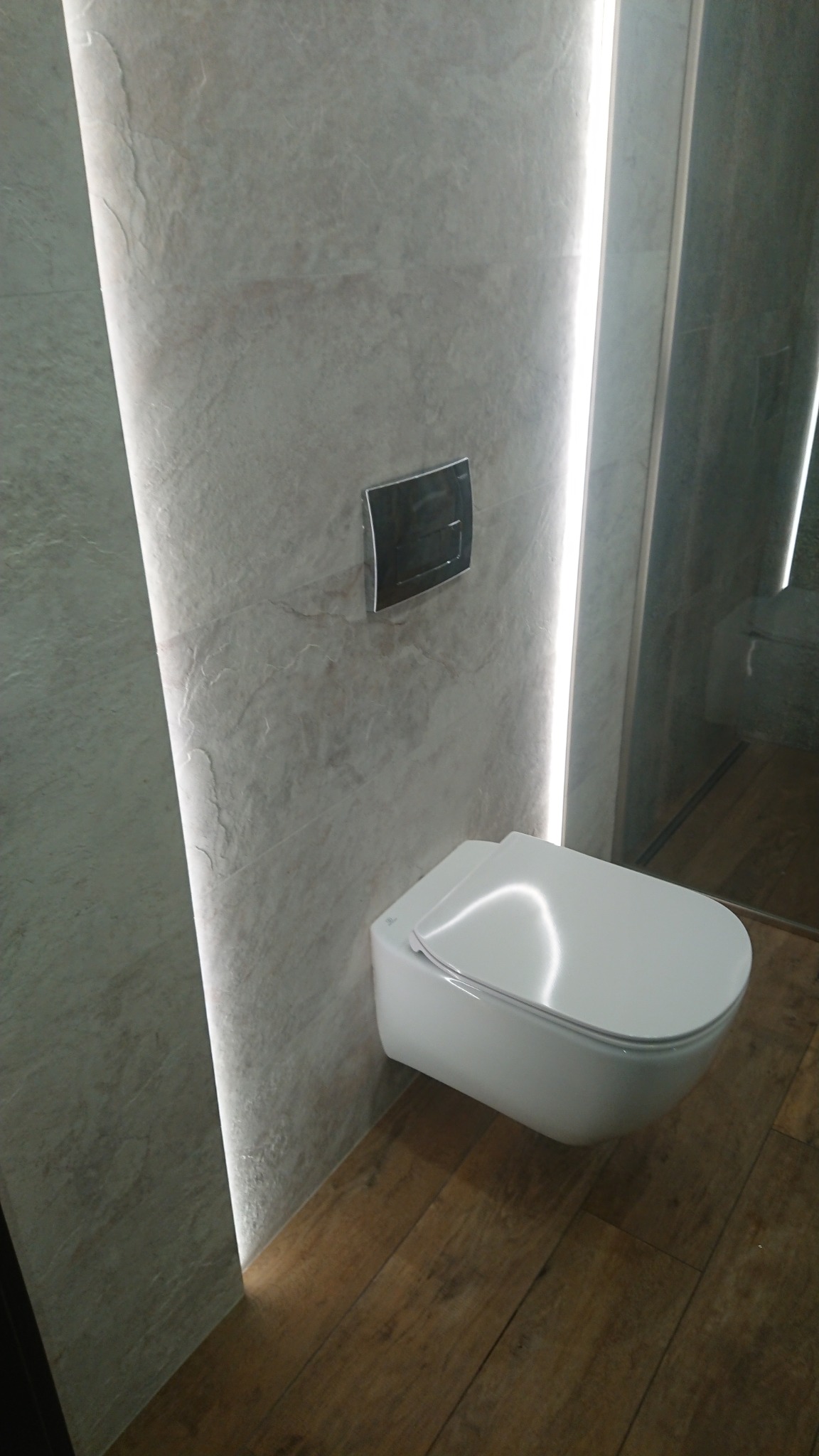 Nowoczesna toaleta podwieszana z chromowanym przyciskiem spłukującym, podświetlana pionowym pasmem LED na ścianie z kamiennej okładziny, na podłodze panele imitujące drewno.