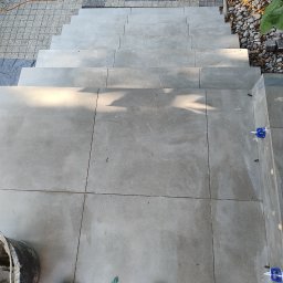 Wiesiek Jamniuk - Wykończone schody zewnętrzne z szarych płytek, widok z góry. Na dole kadru widoczne narzędzia glazurnicze i buty robocze, co sugeruje trwające prace wykończeniowe.