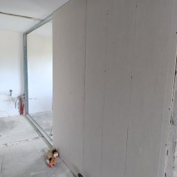 Wiesiek Jamniuk - Montaż ścianki działowej z płyt gipsowo-kartonowych. Widoczne profile, płyty, wkrętarka i karton po wkrętach. W tle instalacja wodna.