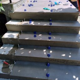 Wiesiek Jamniuk - Układanie płytek na schodach zewnętrznych z użyciem systemu poziomowania; widoczne narzędzia glazurnicze i fragment osoby pracującej na górnym stopniu.