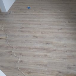 Wiesiek Jamniuk - Ułożone panele podłogowe w pokoju, widoczne listwy przypodłogowe i kabel. Nowa podłoga z jasnego drewna z widoczną strukturą.