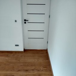 Wiesiek Jamniuk - Białe drzwi z czarnymi paskami na tle białej ściany i drewnianej podłogi. Widoczne gniazdka elektryczne. Minimalistyczne wykończenie wnętrza.