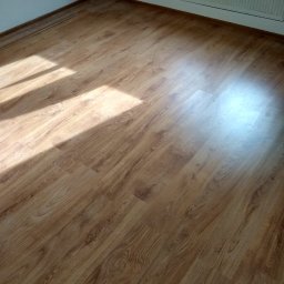 Wiesiek Jamniuk - Jasne panele podłogowe z widocznym wzorem drewna, oświetlone naturalnym światłem słonecznym w pustym pokoju z białymi ścianami i grzejnikiem.