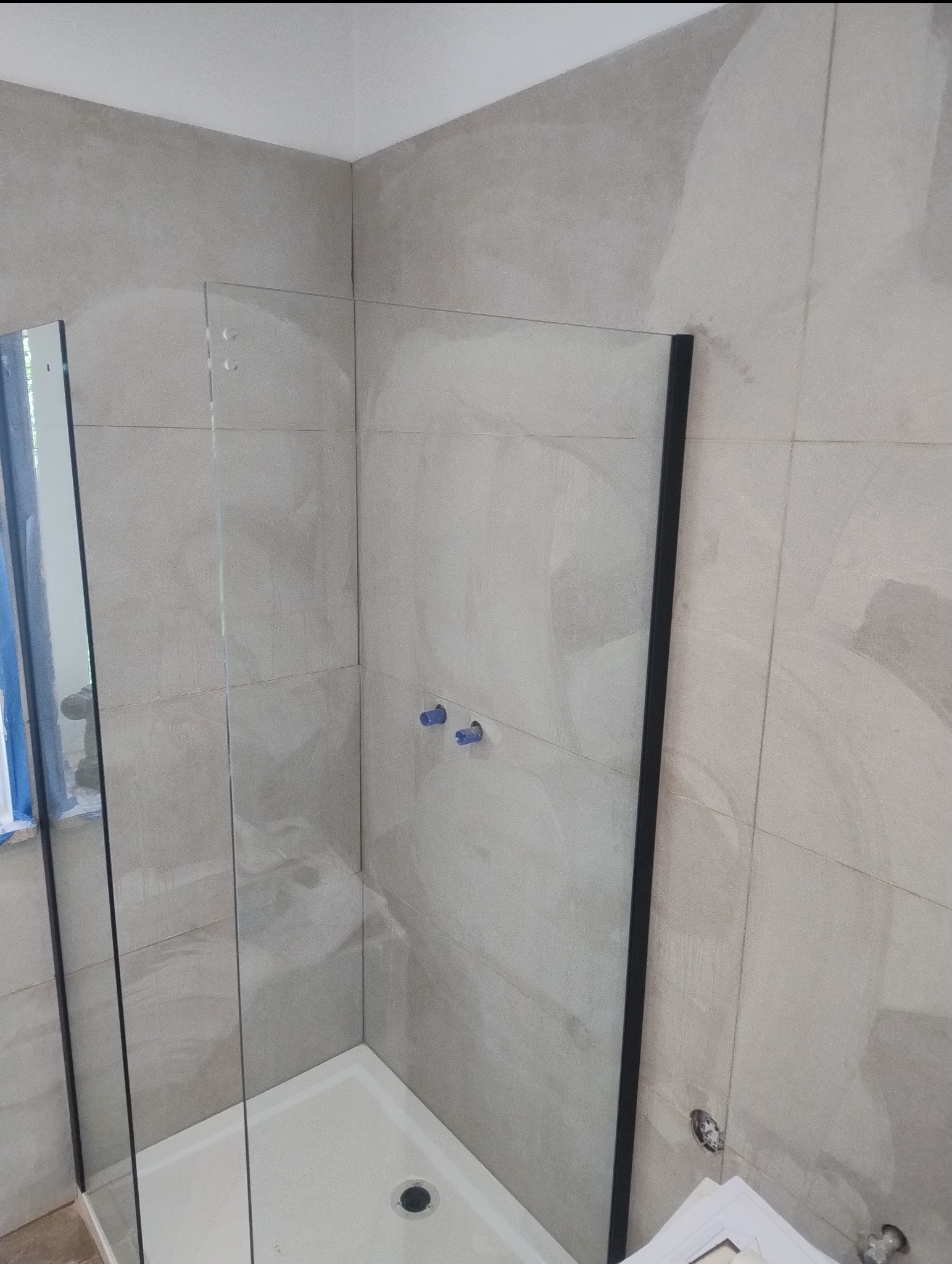 Wykończona kabina prysznicowa z szklaną ścianką i beżowymi płytkami. Widoczne niebieskie zaślepki na wystających rurach. Minimalistyczny design.