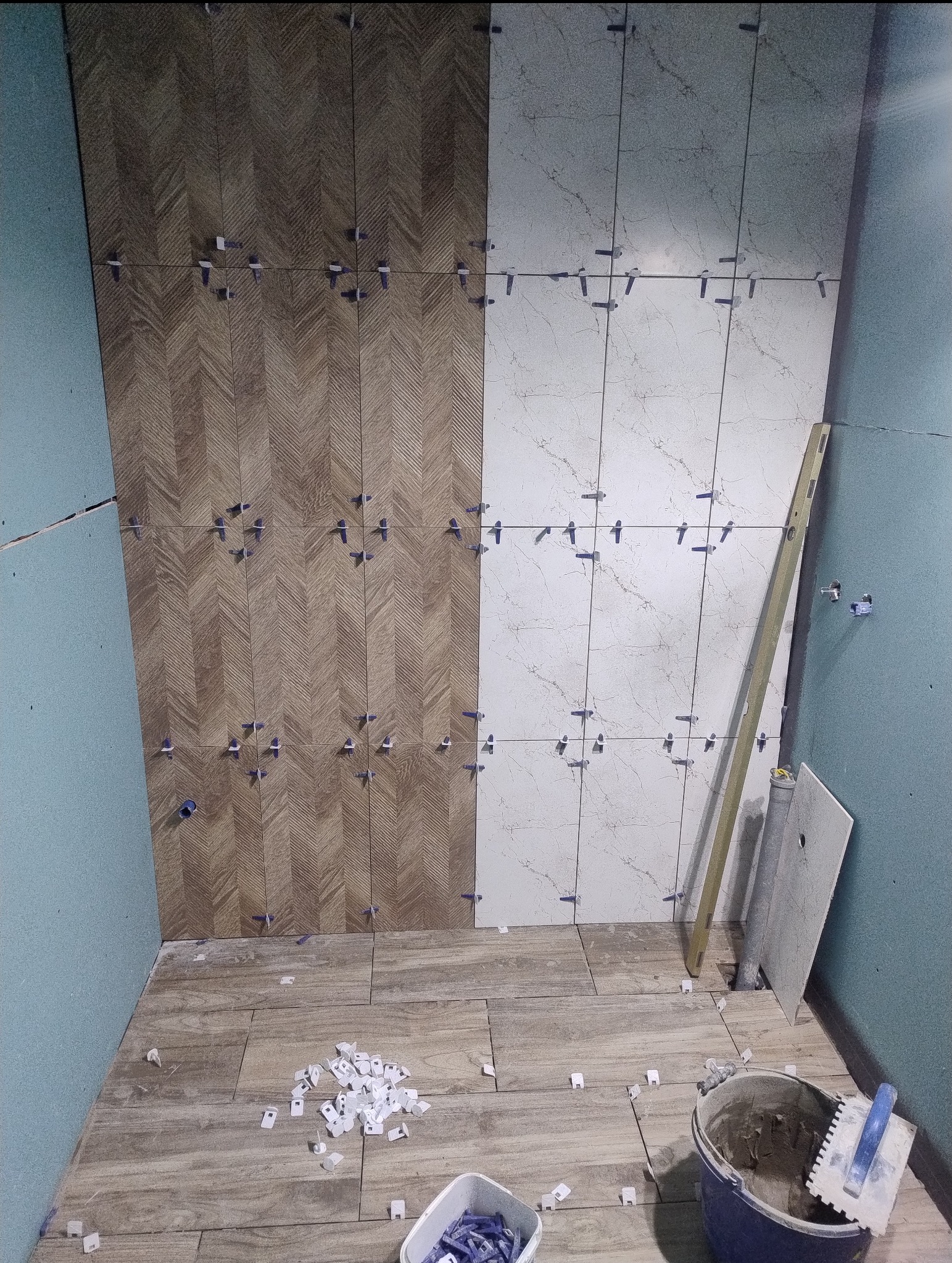 Układanie płytek w łazience: część ściany wykończona w jodełkę, druga część w jasnym marmurze. Widoczne poziomica i narzędzia glazurnicze.