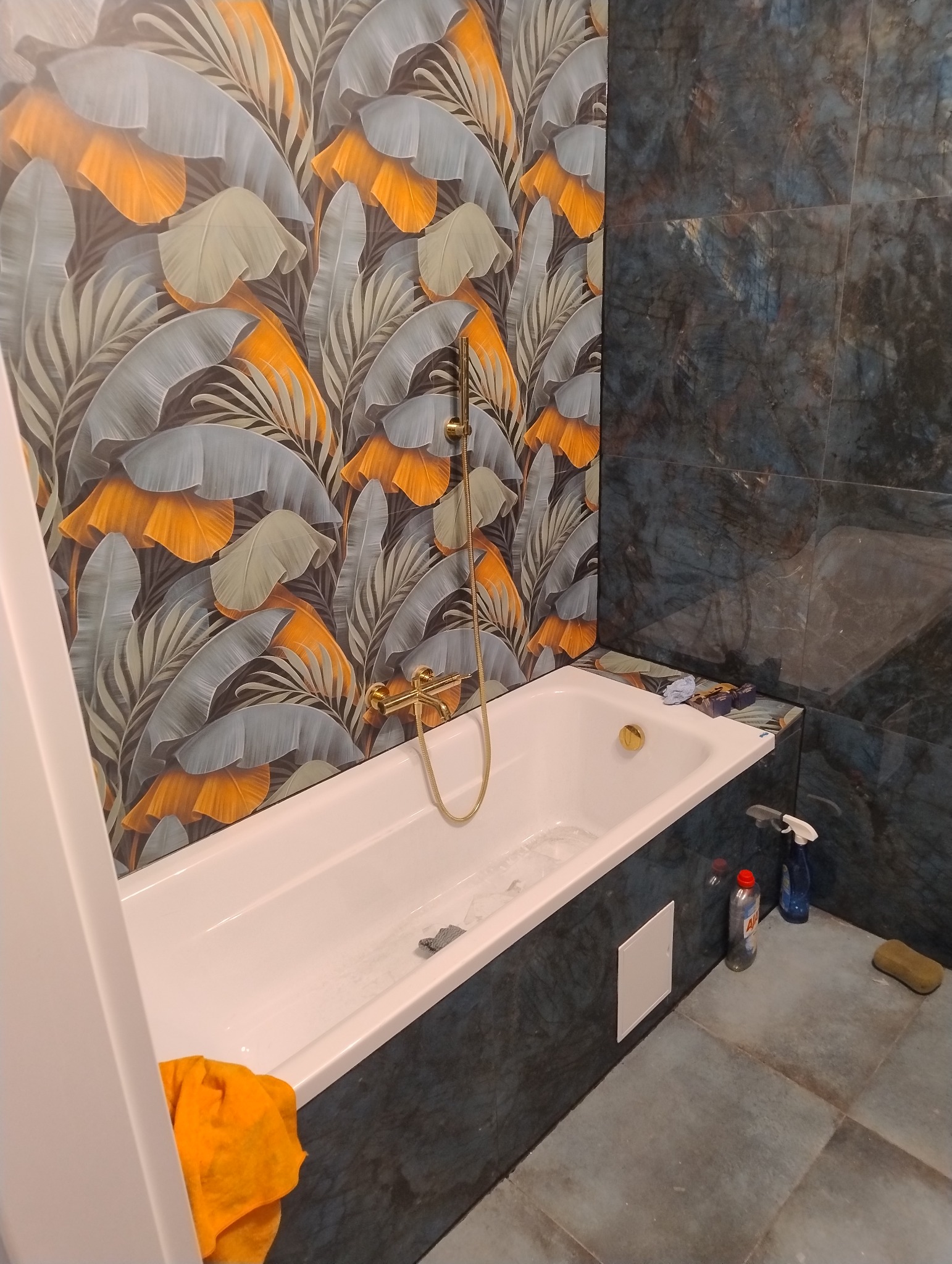 Wykończona łazienka z wanną obudowaną ciemnym gresem. Na ścianie tapeta w tropikalne liście, złota armatura. Widoczne środki czystości.