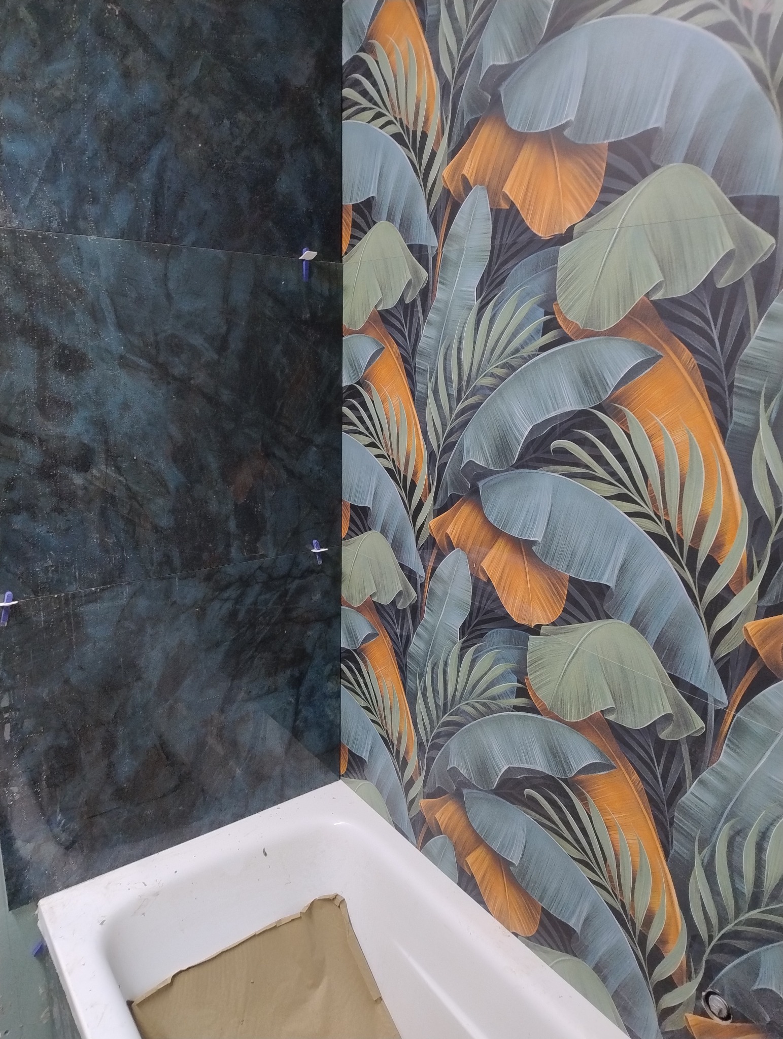 Układanie płytek w łazience: część ściany z ciemnymi płytkami imitującymi kamień, część z wzorem liści. Widoczna wanna zabezpieczona kartonem.