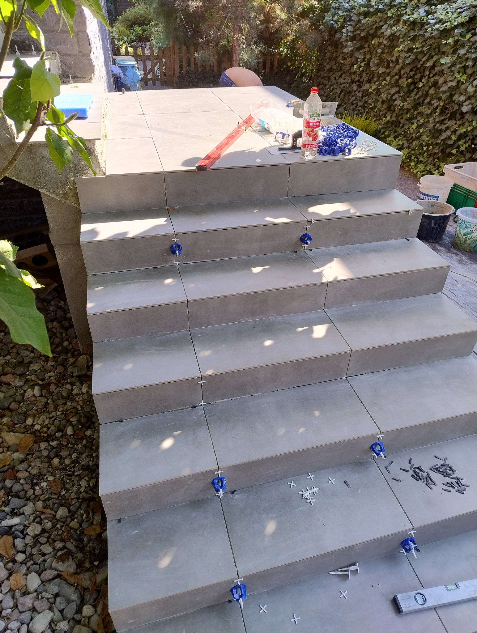 Schody wyłożone szarą terakotą z użyciem poziomicy i niebieskich krzyżyków dystansowych. Widoczne narzędzia i akcesoria glazurnicze, tło z zielenią.