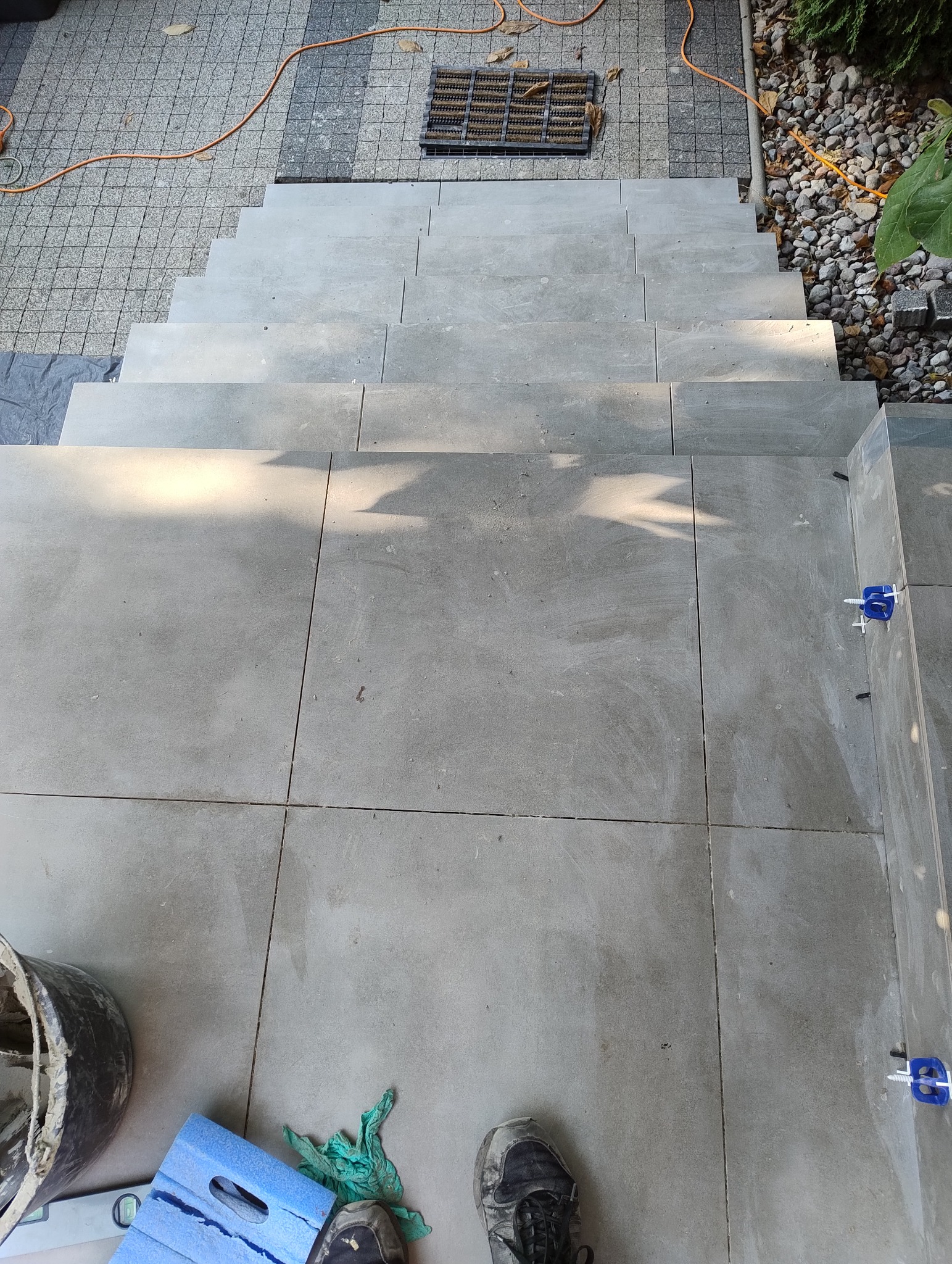 Wykończone schody zewnętrzne z szarych płytek, widok z góry. Na dole kadru widoczne narzędzia glazurnicze i buty robocze, co sugeruje trwające prace wykończeniowe.