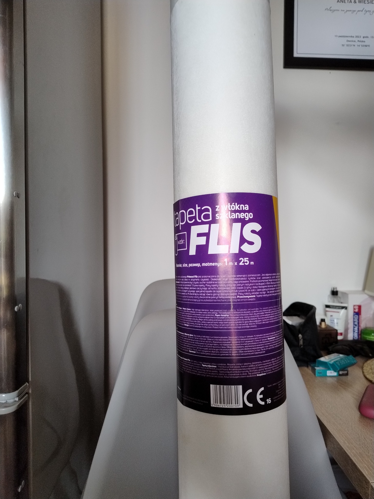 Rolka tapety z włókna szklanego FLIS, z widoczną etykietą informacyjną o rozmiarze 1m x 25m, ustawiona pionowo. Tło stanowią elementy wyposażenia wnętrza.