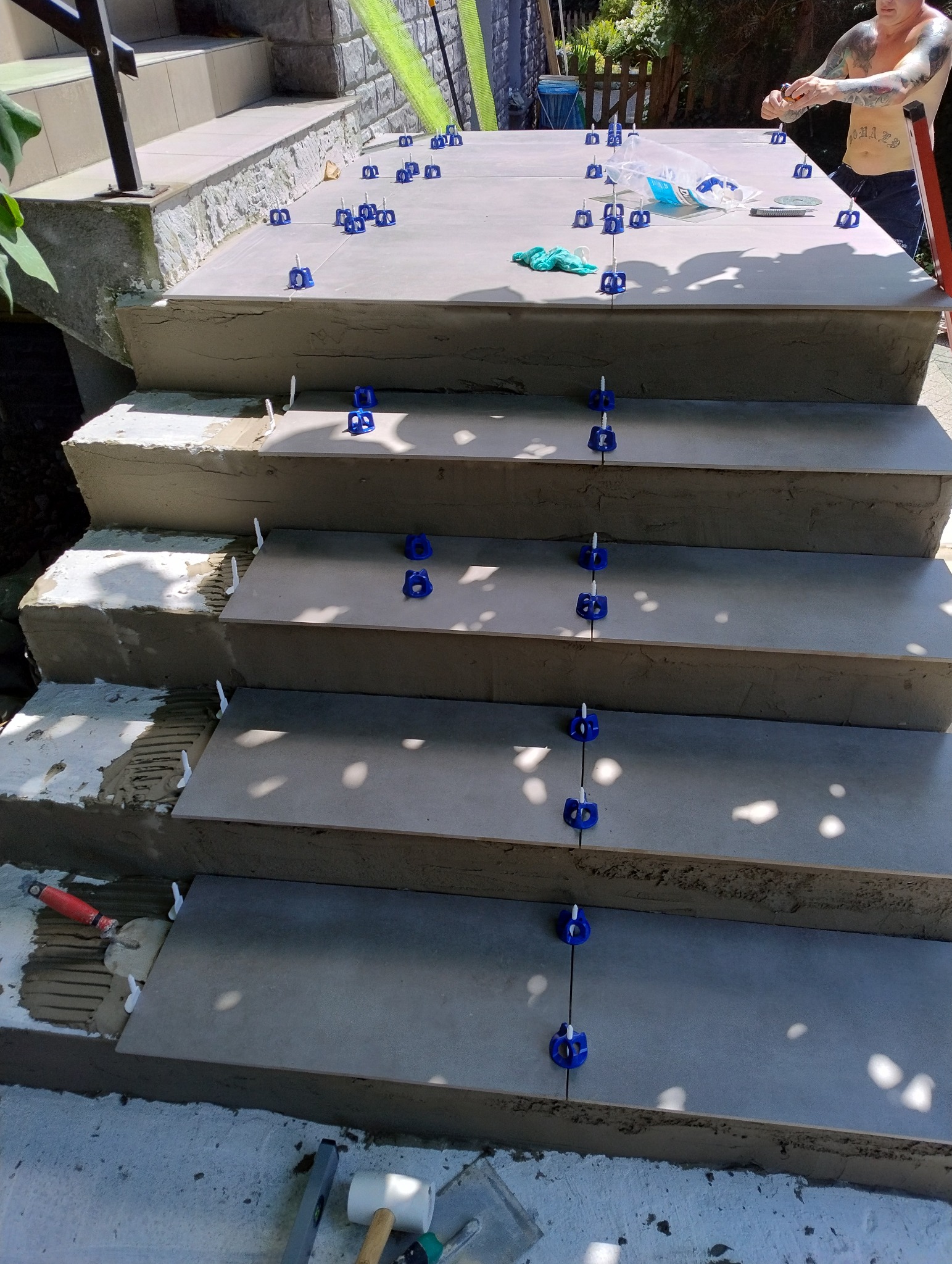 Układanie płytek na schodach zewnętrznych z użyciem systemu poziomowania; widoczne narzędzia glazurnicze i fragment osoby pracującej na górnym stopniu.