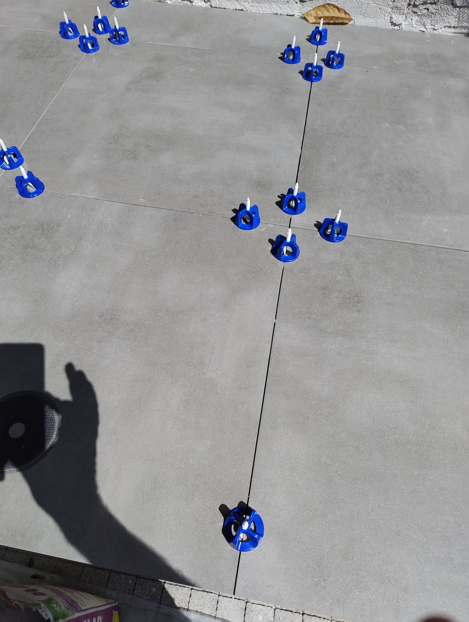 Szare płytki podłogowe z niebieskimi klipsami poziomującymi i cienkimi fugami, widok z góry, cień osoby w lewym dolnym rogu. Układanie płytek z systemem poziomowania.