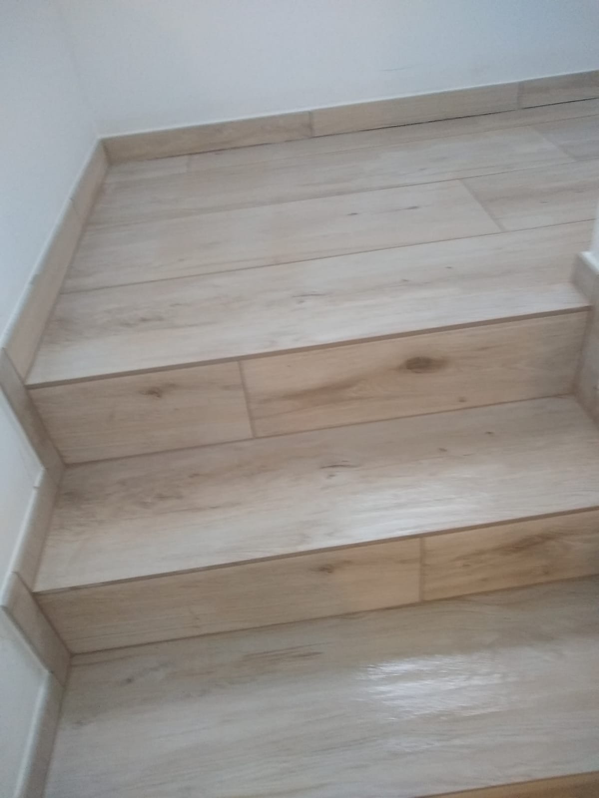 Schody wykończone jasnymi płytkami imitującymi drewno, z delikatnym wzorem słojów. Widoczne precyzyjne ułożenie płytek i estetyczne wykończenie krawędzi.