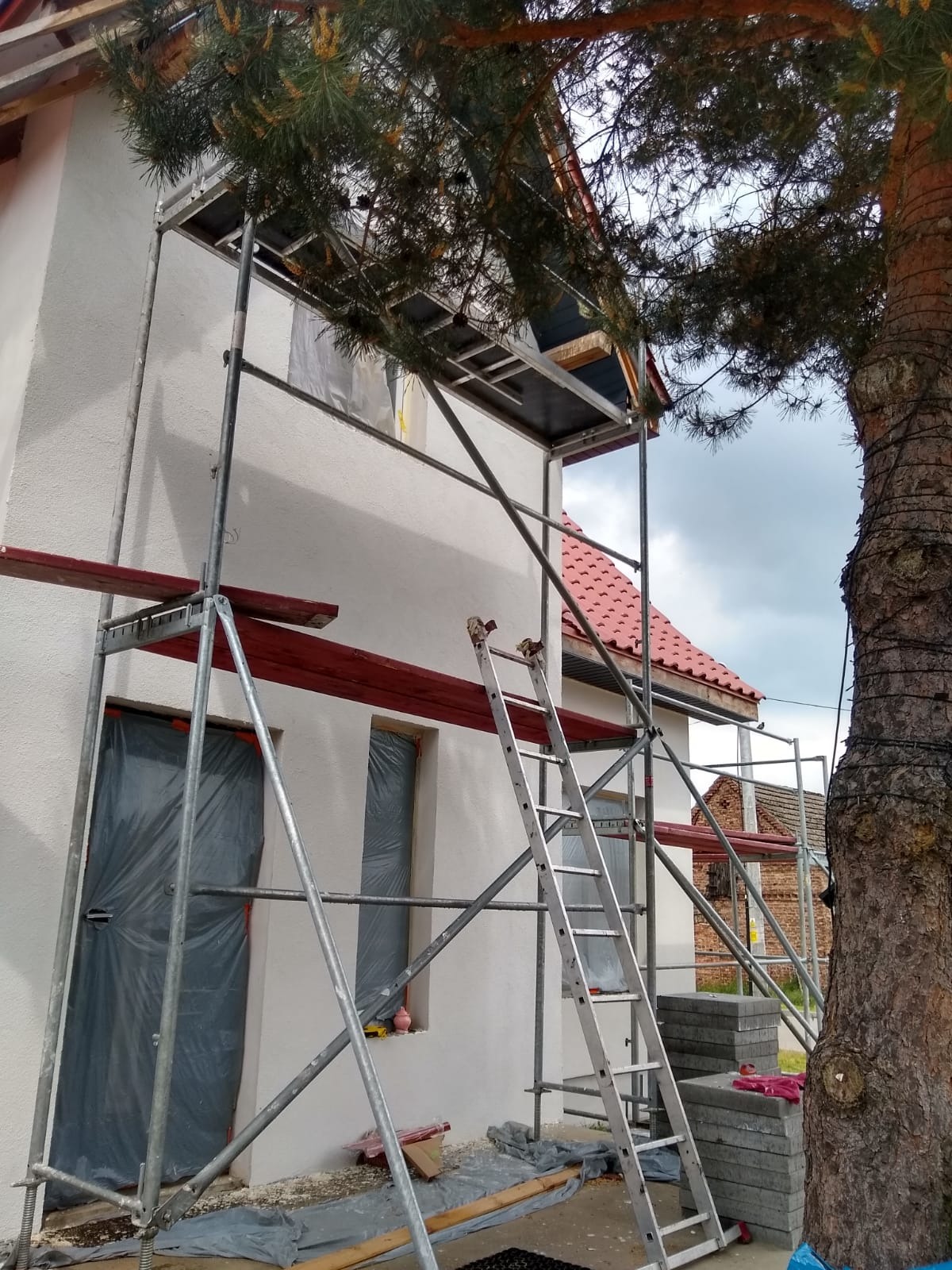 Ściana domu w trakcie prac wykończeniowych, zabezpieczone okna i drzwi folią, rusztowanie, widoczna część dachu z czerwoną dachówką, drzewo iglaste.