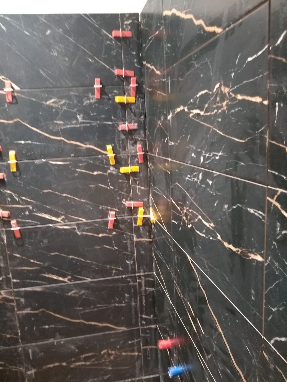 Ściana z czarnych płytek imitujących marmur, z użyciem czerwonych i żółtych krzyżyków dystansowych, w trakcie układania. Widok z bliska na narożnik.