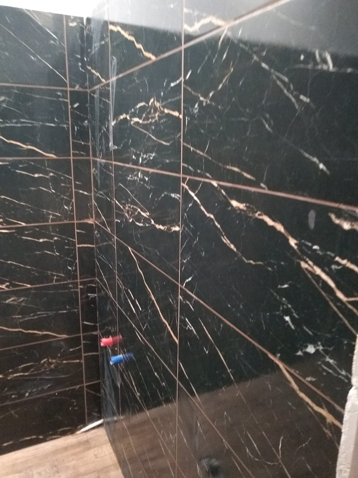 Wykończona ściana w łazience z czarnych, marmurkowych płytek z złotymi żyłkami. Widoczne fragmenty podłogi i przyłącza wodne.