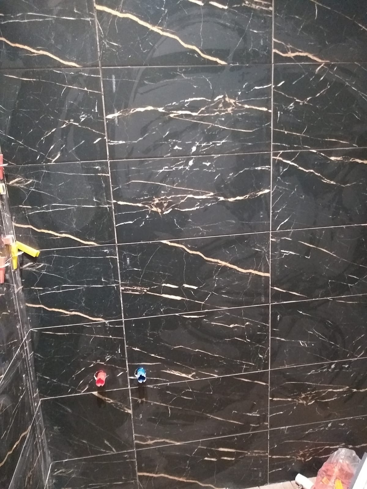 Ułożone, czarne płytki imitujące marmur z wyraźnym, złotym użyleniem. Widoczne krzyżyki dystansowe i wystające rury instalacji wodnej. Świeżo położona glazura.