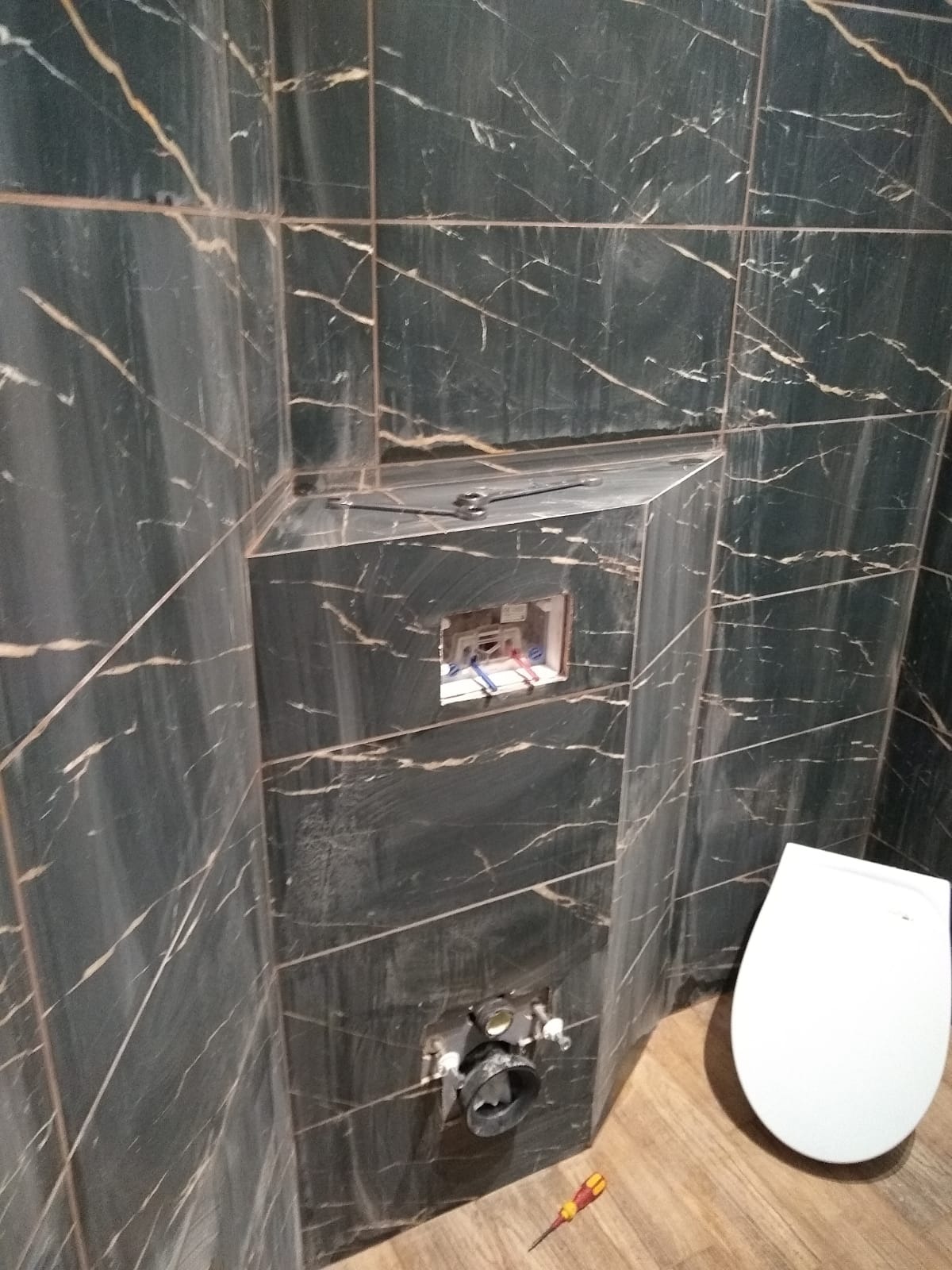 Wykończenie łazienki: czarne płytki z marmurowym wzorem, widoczna instalacja podtynkowego WC, biała miska WC i narzędzie na podłodze.