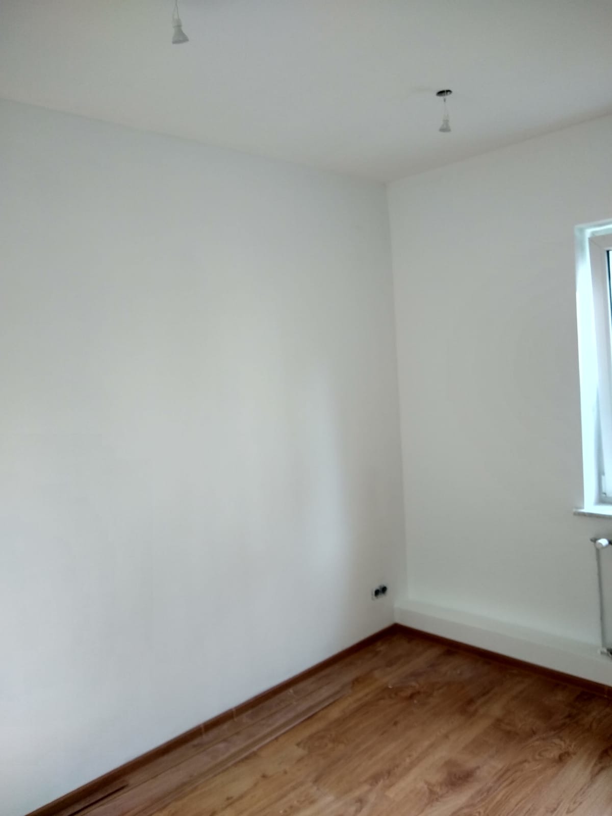 Pomalowane na biało ściany w rogu pokoju z drewnianą podłogą, widoczne gniazdka elektryczne i grzejnik pod oknem. Sufit z dwoma punktami oświetleniowymi.