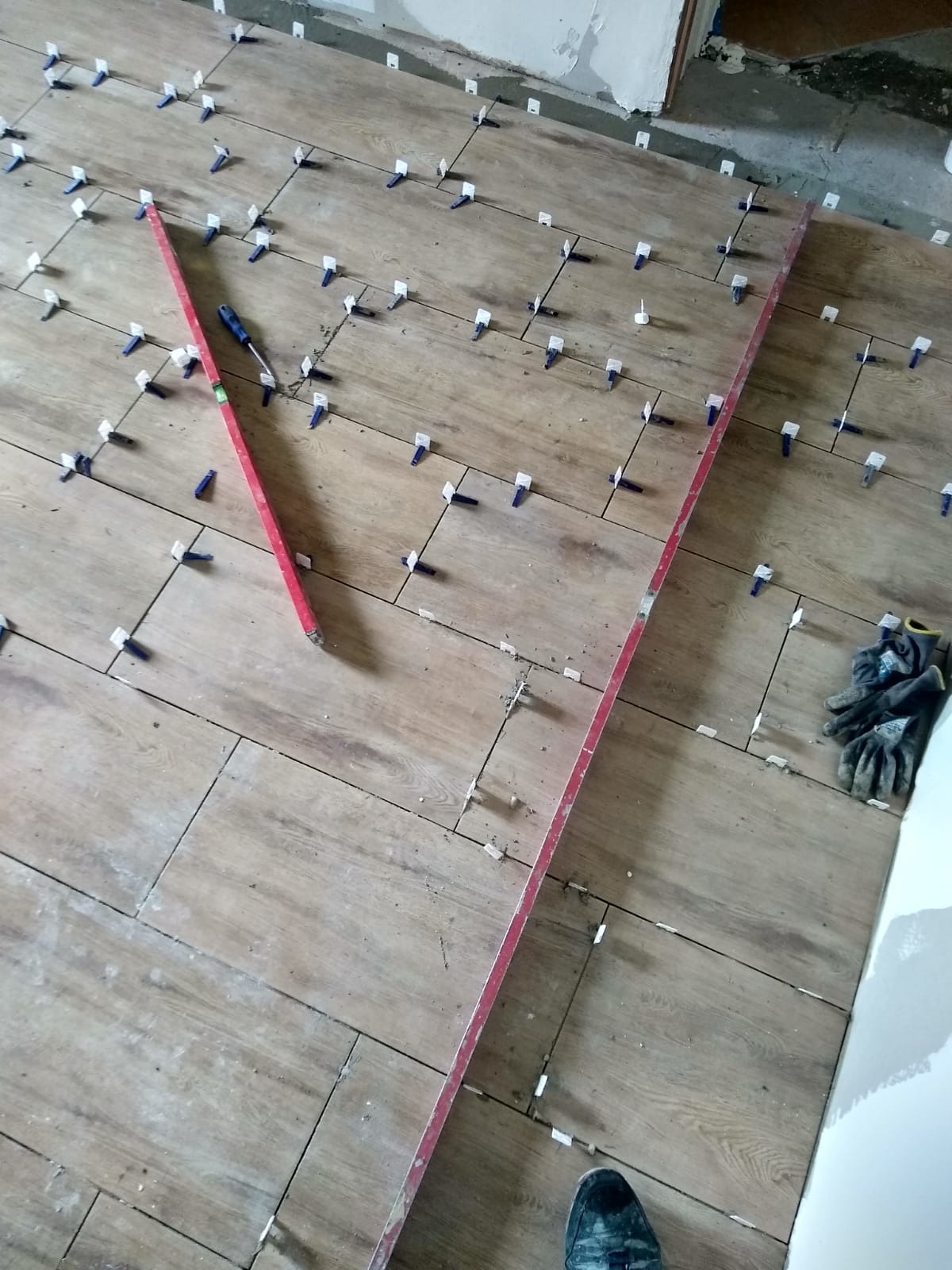 Układanie drewnopodobnych płytek podłogowych z użyciem systemu poziomowania. Widoczne poziomice, śrubokręt i rękawice robocze. Częściowo ukończona podłoga w trakcie prac.