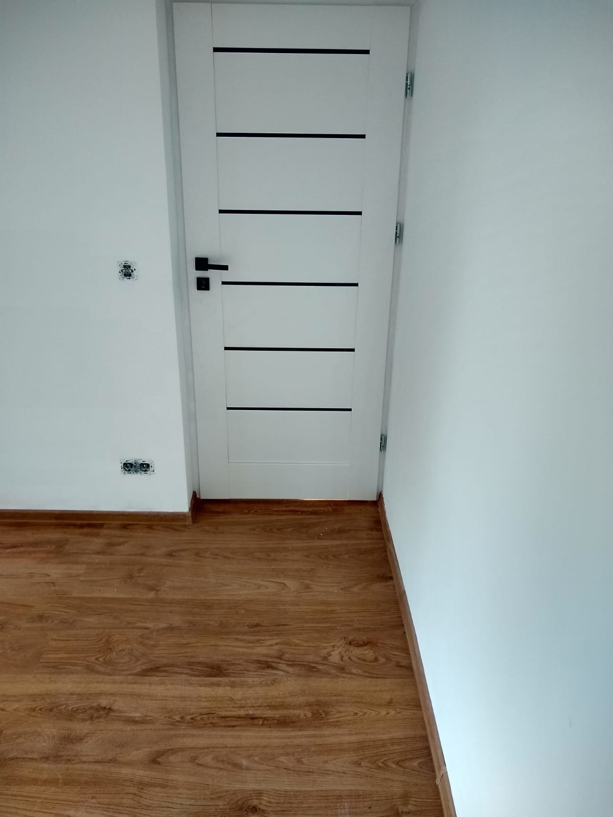 Białe drzwi z czarnymi paskami na tle białej ściany i drewnianej podłogi. Widoczne gniazdka elektryczne. Minimalistyczne wykończenie wnętrza.