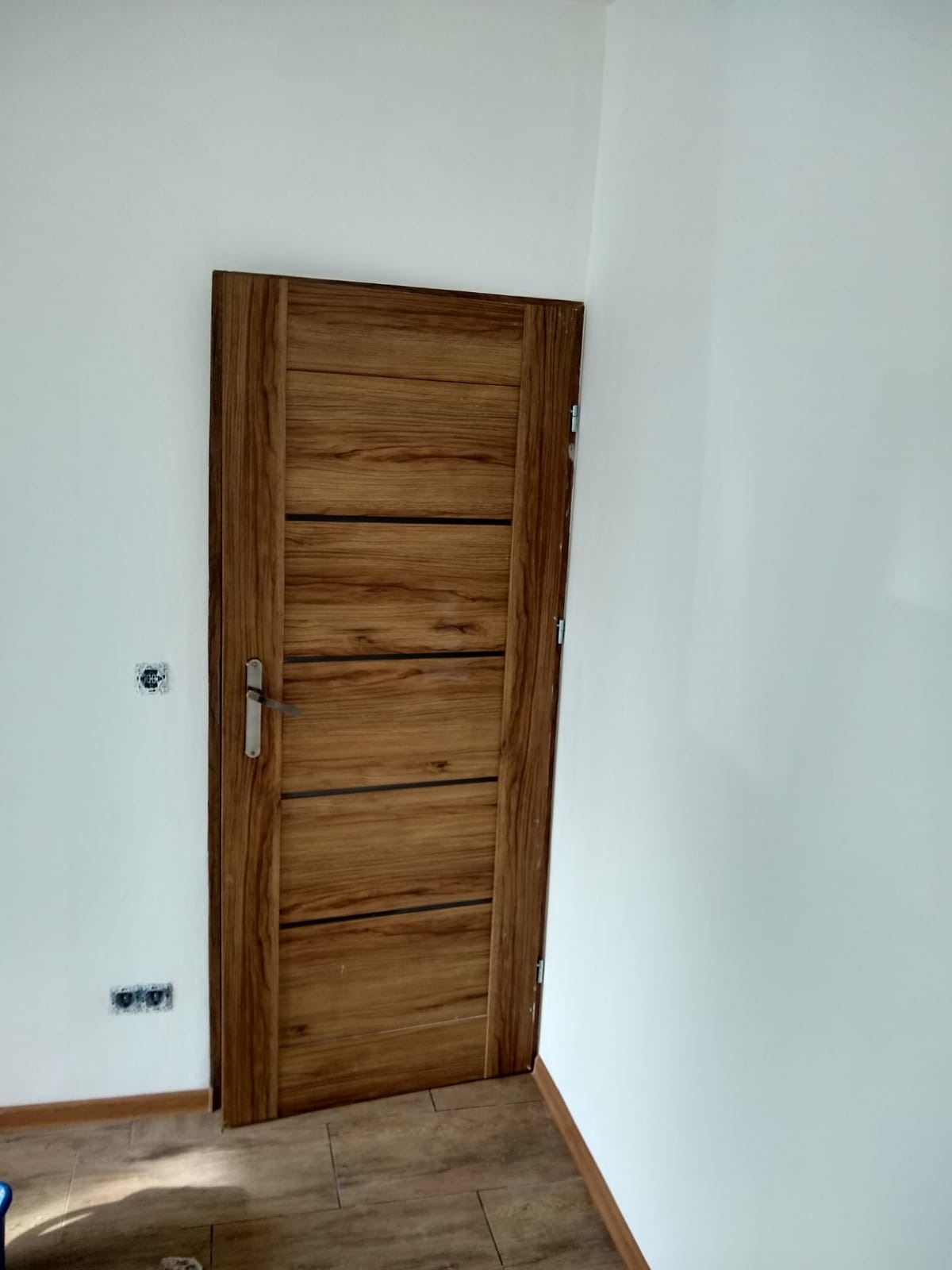 Zamknięte, drewniane drzwi wewnętrzne z poziomymi, czarnymi paskami na tle białej ściany z widocznymi gniazdkami elektrycznymi i brązową podłogą.