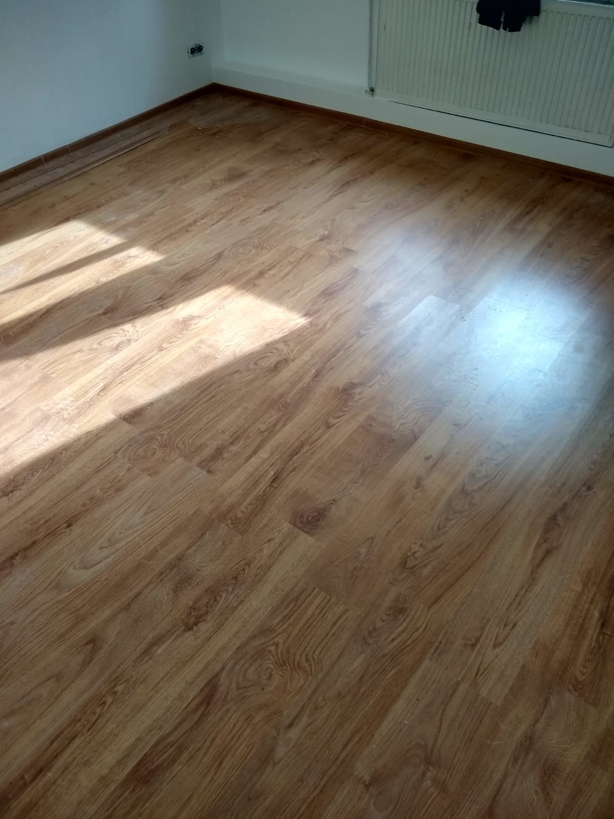 Jasne panele podłogowe z widocznym wzorem drewna, oświetlone naturalnym światłem słonecznym w pustym pokoju z białymi ścianami i grzejnikiem.