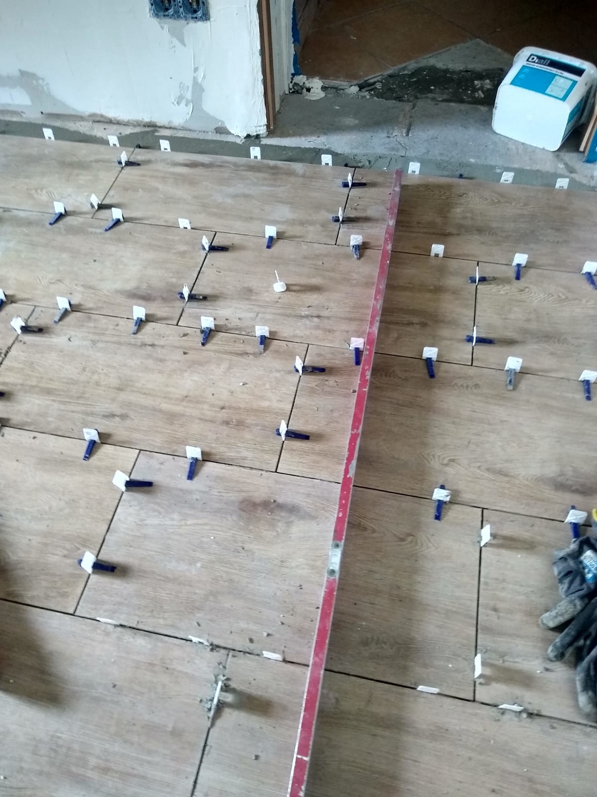 Układanie płytek podłogowych imitujących drewno z użyciem systemu poziomowania. Widoczna poziomica i zapas płytek w tle. Prace glazurnicze w trakcie.