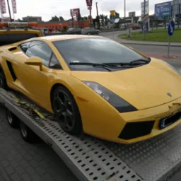 Żółte Lamborghini Gallardo z uszkodzonym przodem, zabezpieczone pasami na platformie lawety w dzień, w tle budynki restauracji fast food.