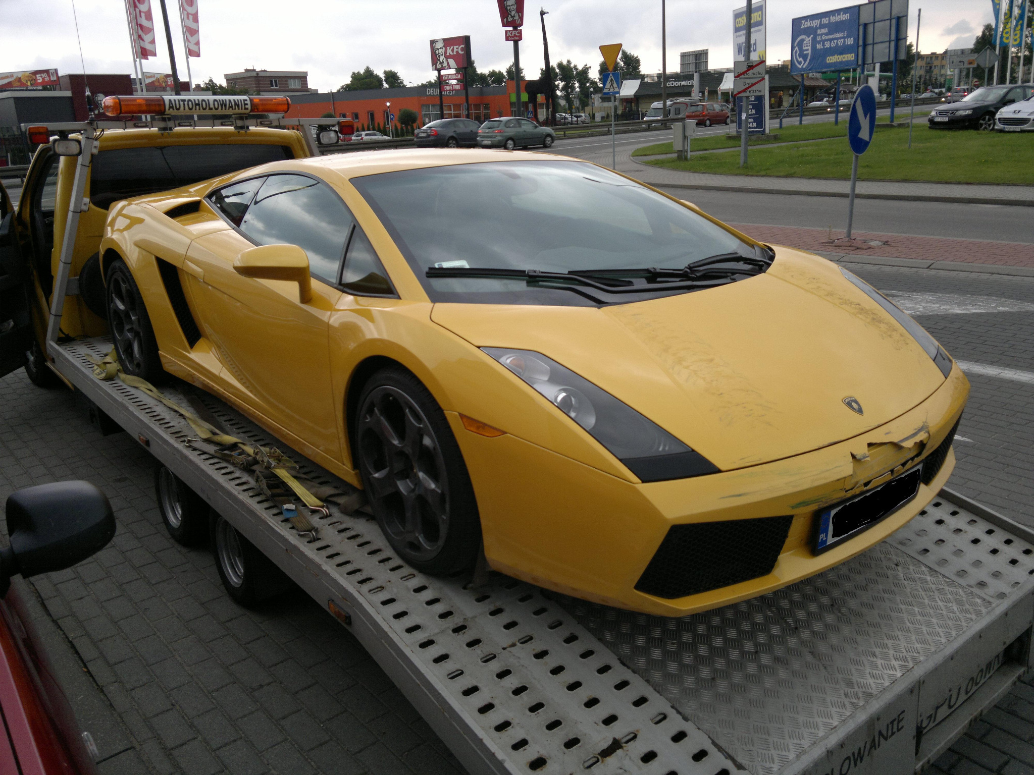 Żółte Lamborghini Gallardo z uszkodzonym przodem, zabezpieczone pasami na platformie lawety w dzień, w tle budynki restauracji fast food.
