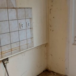 ITD. Usługi Budowlane - Kąt w kuchni w trakcie demontażu: stare płytki częściowo usunięte, odsłonięte gniazdka elektryczne, resztki kleju na ścianach, gruz na podłodze z płytek.
