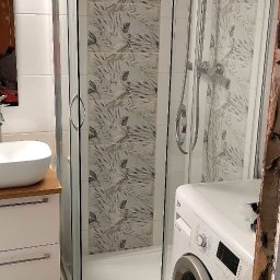 ITD. Usługi Budowlane - Wykończona łazienka z kabiną prysznicową z deszczownicą, umywalką na szafce i pralką. Na ścianach płytki z motywem liści. Widoczny fragment ściany w trakcie remontu.