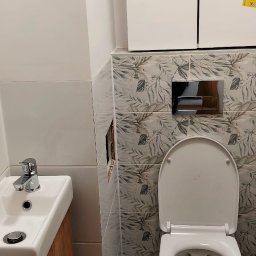 ITD. Usługi Budowlane - Wykończona łazienka: biała umywalka z drewnianą szafką, toaleta z zabudowaną spłuczką i płytki z motywem liści. Na ścianie naklejki informacyjne.