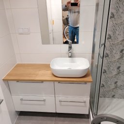 ITD. Usługi Budowlane - Nowoczesna łazienka: biała szafka z drewnianym blatem, umywalka nablatowa i kabina prysznicowa z roślinnym wzorem na płytkach. W lustrze odbicie osoby robiącej zdjęcie.