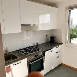 ITD. Usługi Budowlane - Nowoczesna kuchnia z białymi, błyszczącymi szafkami i blatem imitującym marmur, widok z bliska na płytę gazową i piekarnik, okno z widokiem na zieleń w tle.