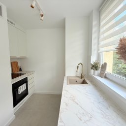 ITD. Usługi Budowlane - Jasna kuchnia z marmurowym blatem, złotą armaturą i dodatkami w stylu boho. Widoczne okno z roletami i dekoracje roślinne. Minimalistyczny design.