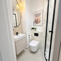 ITD. Usługi Budowlane - Nowoczesna łazienka w beżach: kabina prysznicowa z czarnymi ramami, umywalka z szafką, wisząca toaleta, lustro w czarnej ramie i grafika na ścianie.