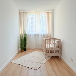 ITD. Usługi Budowlane - Jasne, minimalistyczne wnętrze pokoju dziecięcego w Oławie z drewnianym łóżeczkiem, dywanem i rośliną w doniczce przy oknie z beżowymi zasłonami.